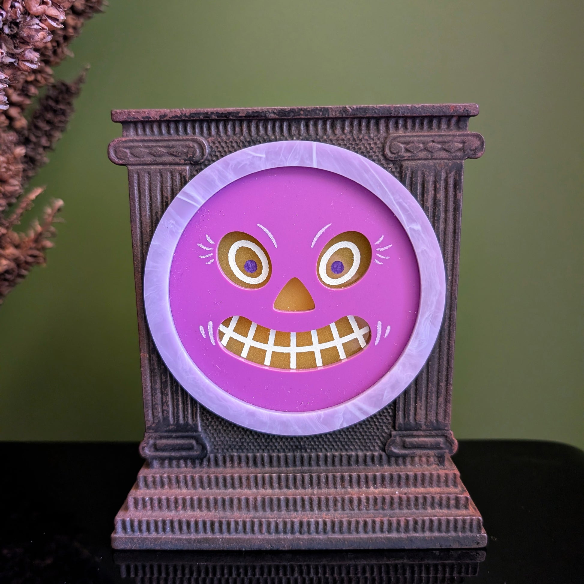 A cast iron clock frame that has a smiling purple jack-o-lantern face is sitting on a black surface with dried mullein behind it. 