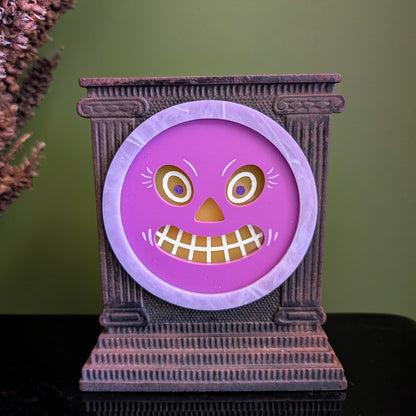 A cast iron clock frame that has a smiling purple jack-o-lantern face is sitting on a black surface with dried mullein behind it. 