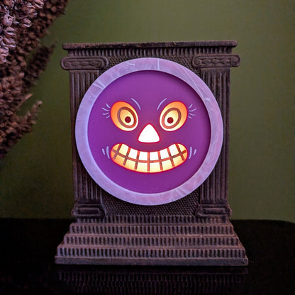 A cast iron clock frame that has a smiling purple jack-o-lantern face is sitting on a black surface with dried mullein behind it. The face is lit up.