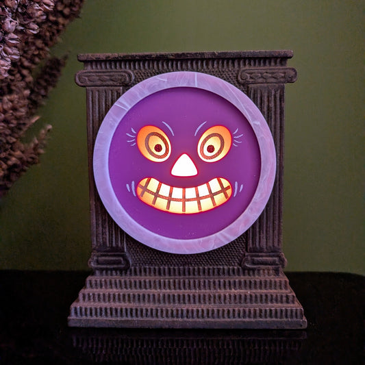 A cast iron clock frame that has a smiling purple jack-o-lantern face is sitting on a black surface with dried mullein behind it. The face is lit up.