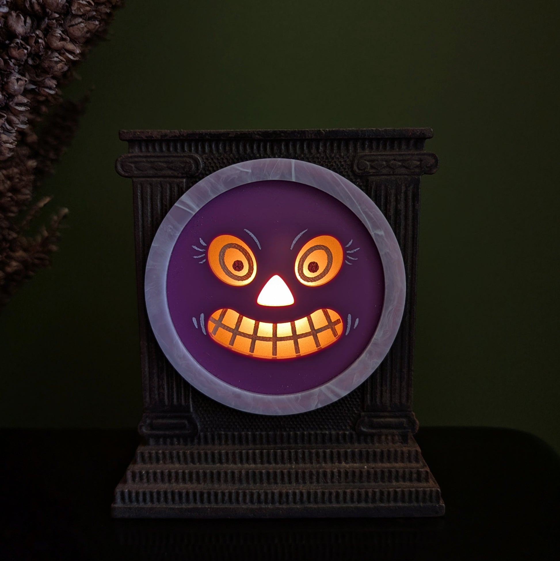 A cast iron clock frame that has a smiling purple jack-o-lantern face is sitting on a black surface with dried mullein behind it. The face is lit up and the background is very dark.