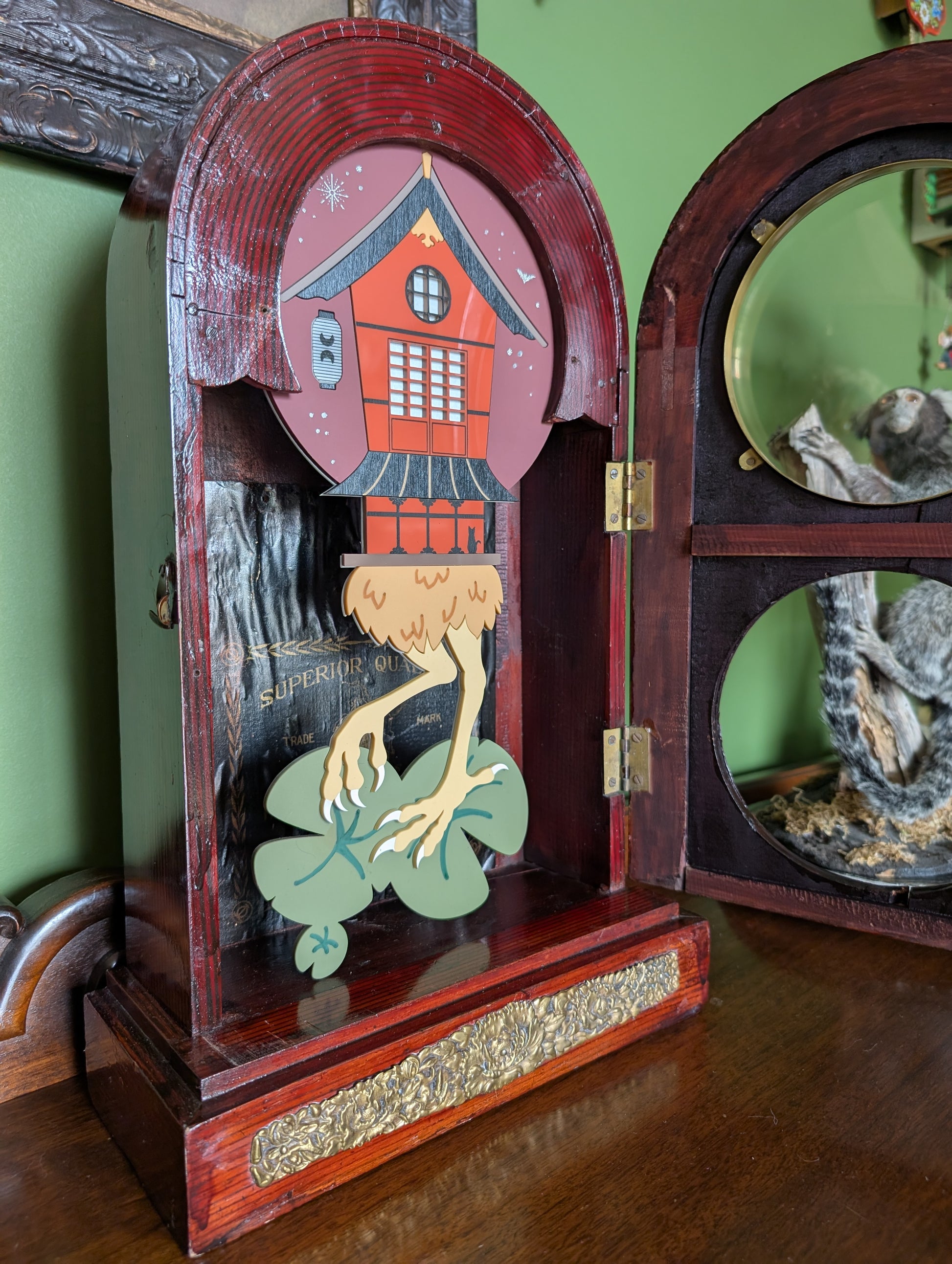 Japanese-inspired Baba Yaga house displays through circular windows of a vintage wooden clock frame. The original clock was made in Japan. The front door is open showing the whole witch house and bird legs standing on lily pads. 