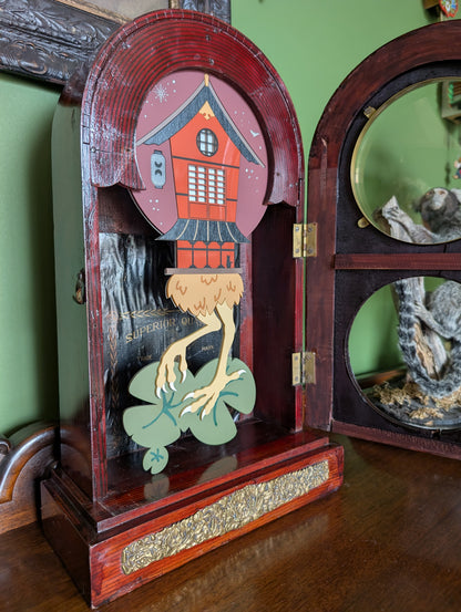 Japanese-inspired Baba Yaga house displays through circular windows of a vintage wooden clock frame. The original clock was made in Japan. The front door is open showing the whole witch house and bird legs standing on lily pads. 