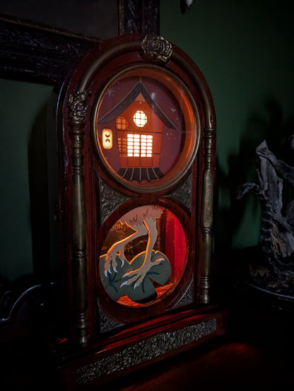 Japanese-inspired Baba Yaga house displays through circular windows of a vintage wooden clock frame. The original clock was made in Japan. The windows and lantern of the house are lit up. The witch scene sits on a table against a green wall in the dark.