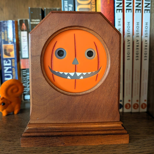 A vintage wood clock frame repurposed with an orange jack-o-lantern face. The clock sits on a book shelf.