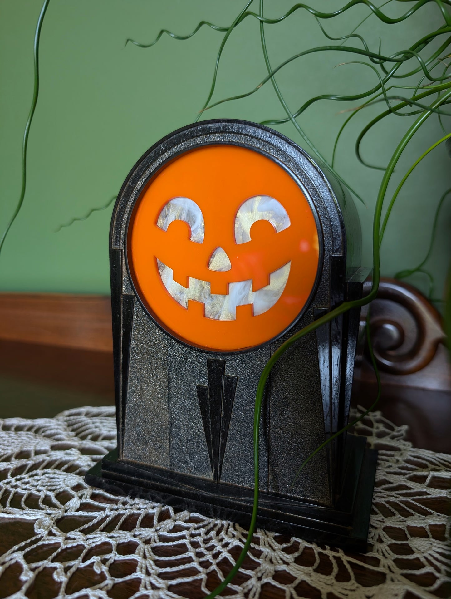 A vintage art deco themed plastic clock frame that has a smiling jack-o-lantern face is sitting on a doily against a green wall.   