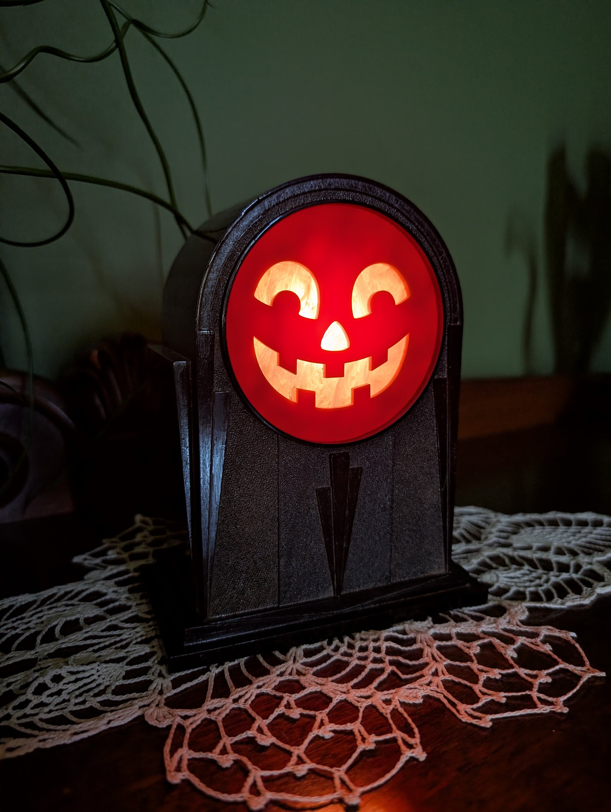 A vintage art deco themed plastic clock frame that has a smiling jack-o-lantern face is sitting on a doily against a green wall.  The scene is dark with the face lit up from behind like Halloween time.
