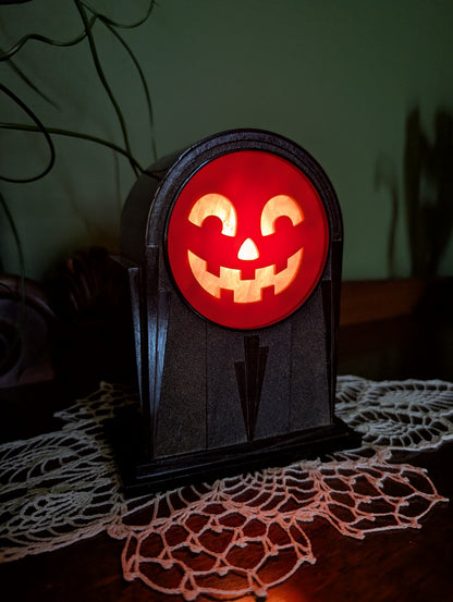 A vintage art deco themed plastic clock frame that has a smiling jack-o-lantern face is sitting on a doily against a green wall.  The scene is dark with the face lit up from behind like Halloween time.