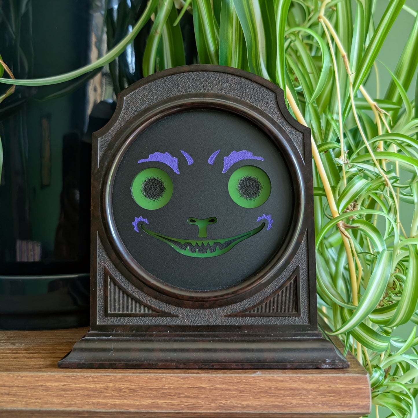 A vintage plastic clock frame that has a black cat jack-o-lantern face is sitting on a shelf with a spider plant behind it. The eyes, nose, and mouth are green.