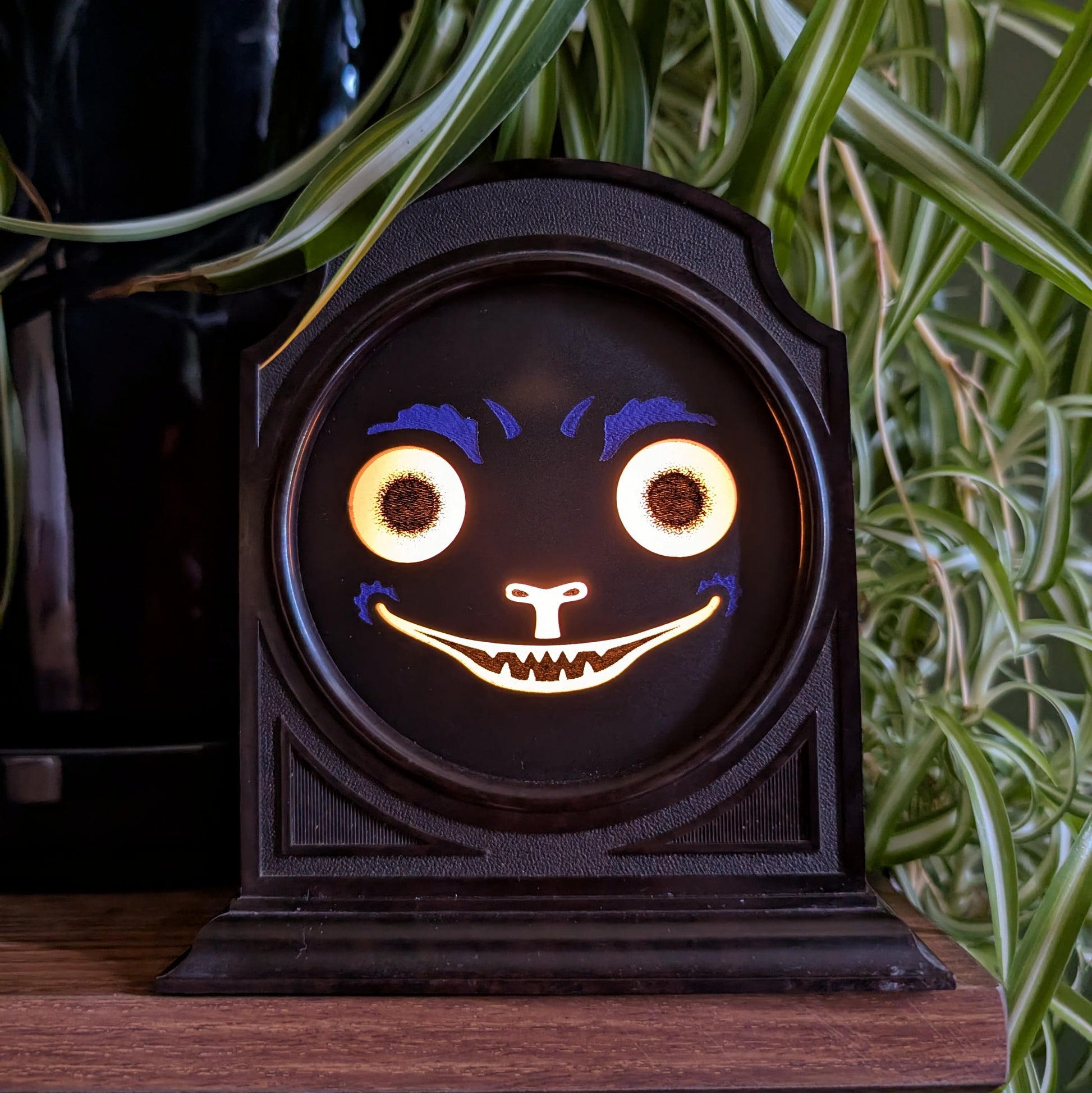 A vintage plastic clock frame that has a black cat jack-o-lantern face is sitting on a shelf with a spider plant behind it. The cat face is lit up.