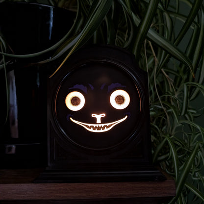 A vintage plastic clock frame that has a black cat jack-o-lantern face is sitting on a shelf with a spider plant behind it. The scene is very dark and cat face is lit up brightly.