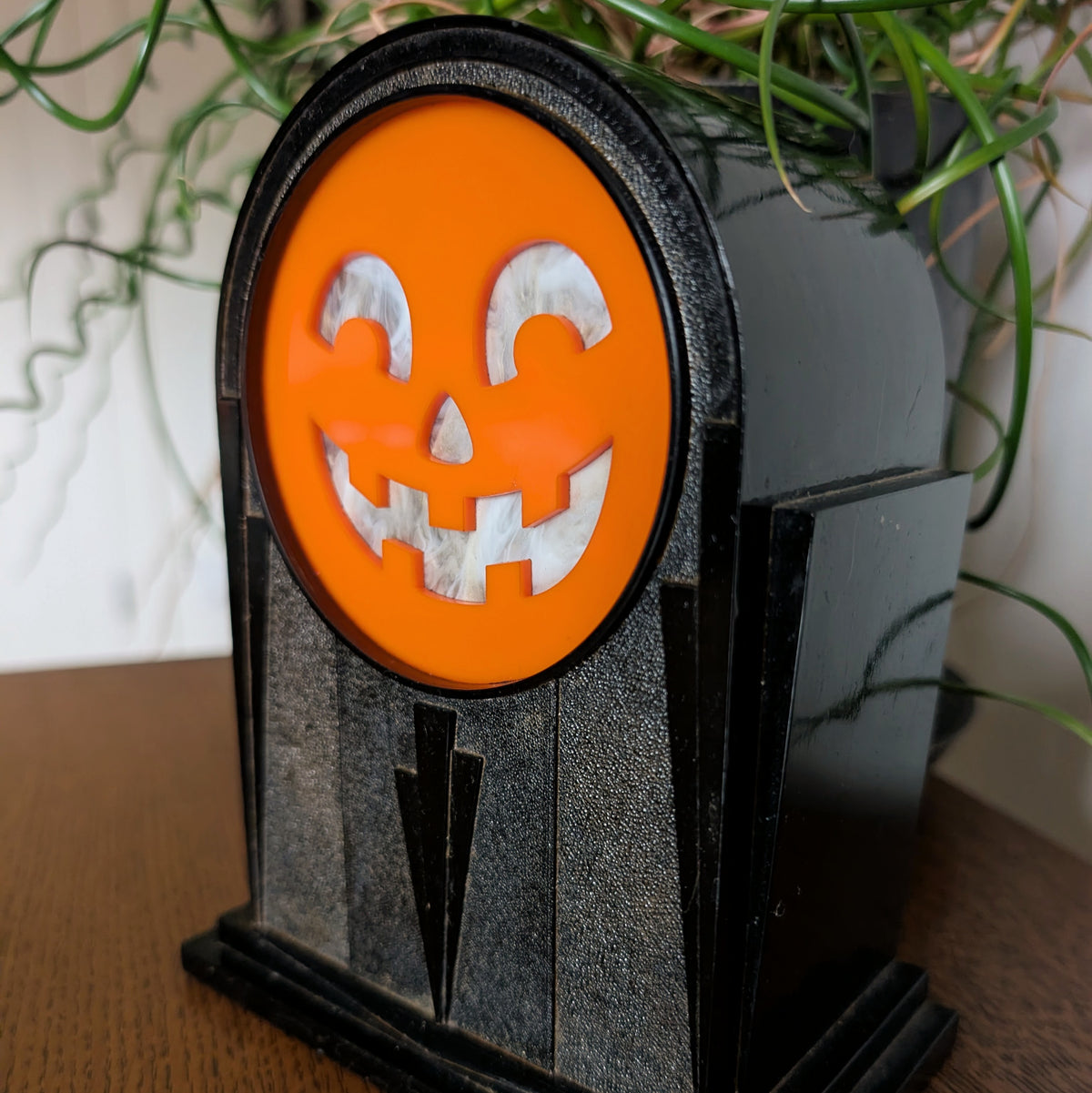 An art deco plastic clock frame features a cheerful acrylic jack-o-lantern face and is sitting on a table against a white and plant filled background.