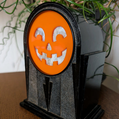 An art deco plastic clock frame features a cheerful acrylic jack-o-lantern face and is sitting on a table against a white and plant filled background.