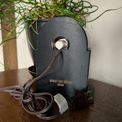 The back of a clock frame with a light and cord attached to the back birch plywood piece. Engraved at the bottom 'ASHLEY HAY DESIGN 2026'. It is sitting on a table against a white and plant filled background. 
