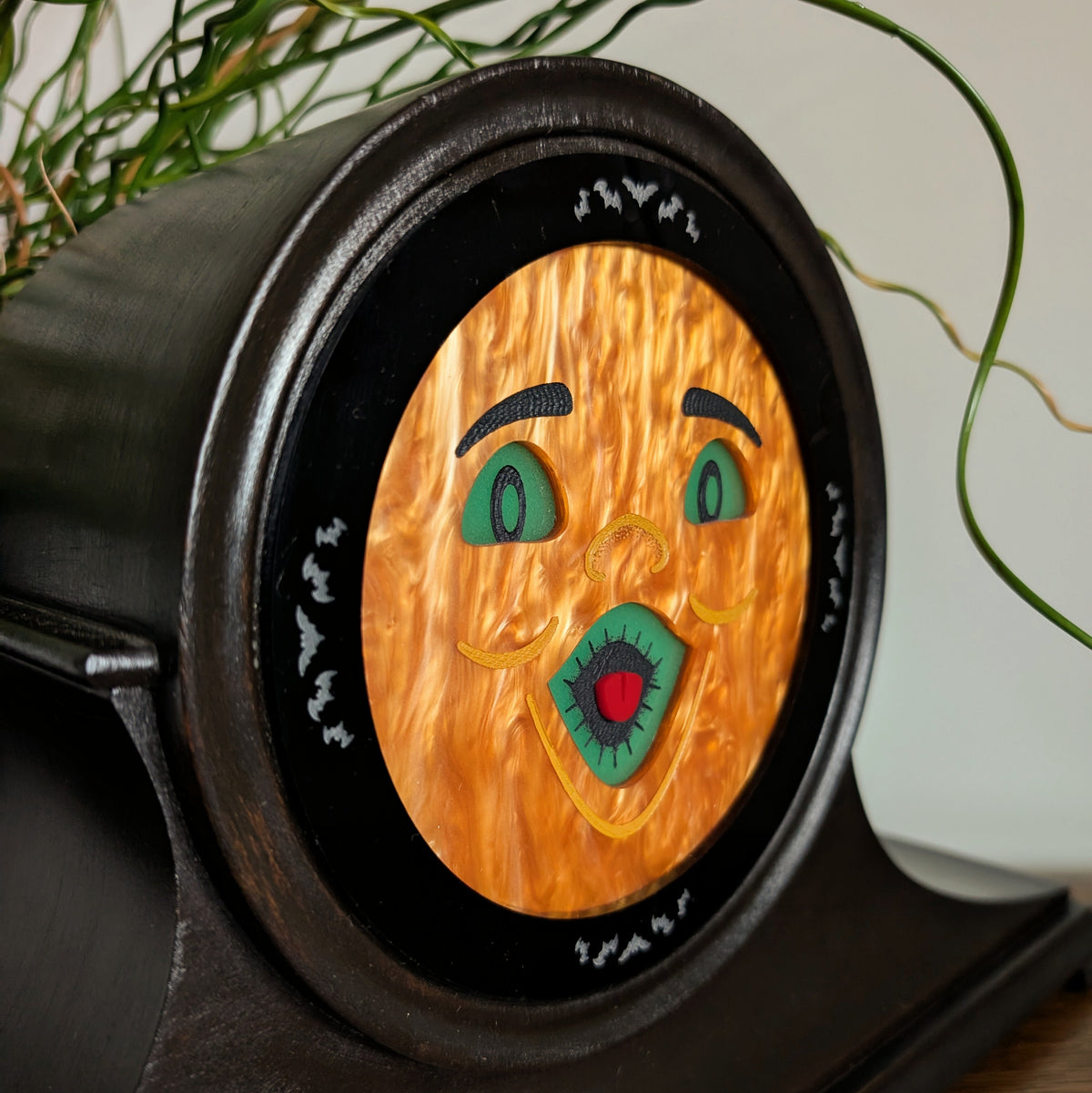 The closeup view of an acrylic jack-o-lantern face inside a vintage wooden clock frame sitting on a table against a white and plant filled background.