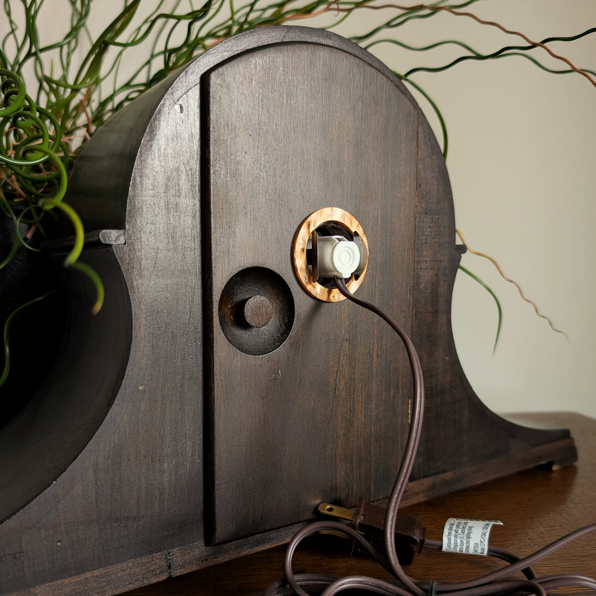 The back of a wooden clock frame with a light and cord attached to the back door. Engraved inside the door reads 'ASHLEY HAY DESIGN 2026'. It is sitting on a table against a white and plant filled background.
