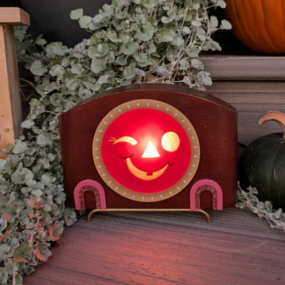 A vintage mid-century modern wooden clock frame that has a winking jack-o-lantern face is sitting on a step against a plants and pumpkins  The scene is dark with the face lit up from inside.