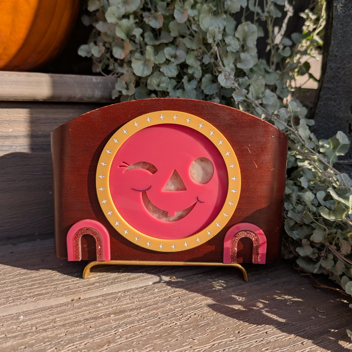 A vintage mid-century modern wooden clock frame that has a winking jack-o-lantern face is sitting on a step against a plants.
