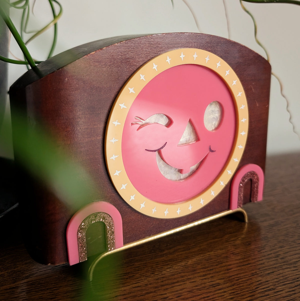 The closeup view of an acrylic winking jack-o-lantern face inside a vintage wooden clock frame sitting on a table against a white and plant filled background.