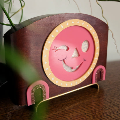 The closeup view of an acrylic winking jack-o-lantern face inside a vintage wooden clock frame sitting on a table against a white and plant filled background.