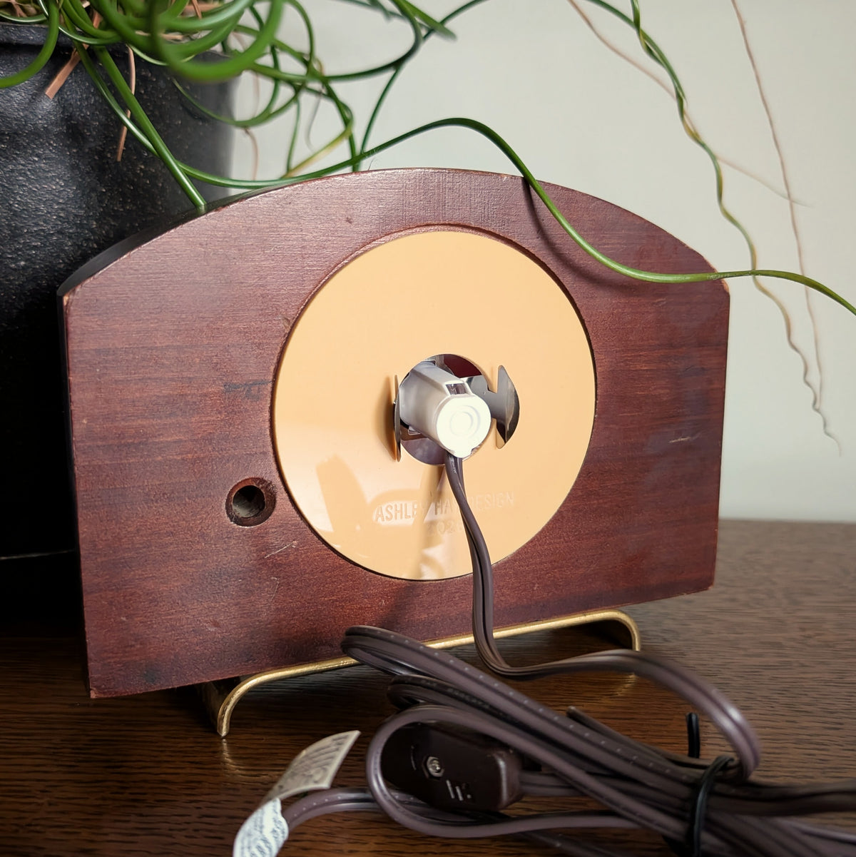 The back of a wooden clock frame with a light and cord attached to the back acrylic piece. Engraved at the bottom 'ASHLEY HAY DESIGN 2026'.  It is sitting on a table against a white and plant filled background.