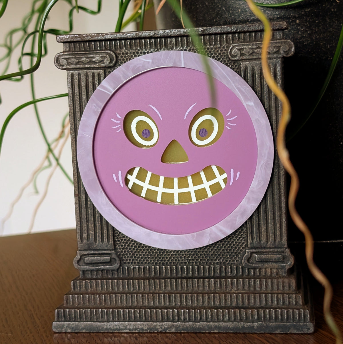 A vintage cast iron clock frame features an acrylic jack-o-lantern face and is sitting on a table against a white and plant filled background.