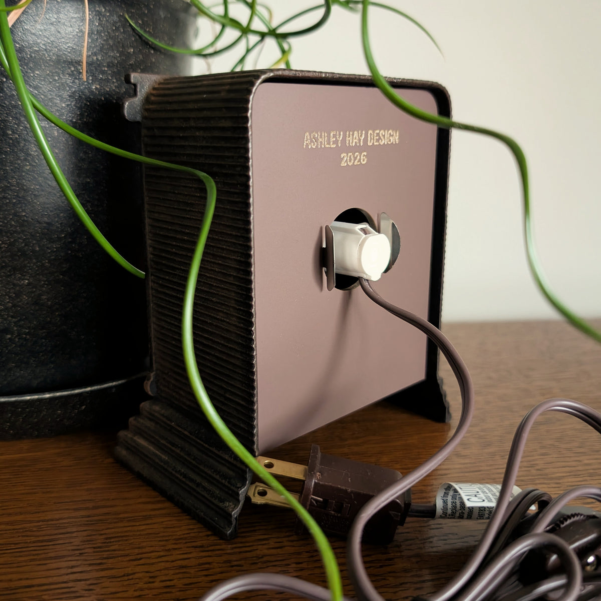 The back of a clock frame with a light and cord attached to the back acrylic piece. Engraved at the bottom 'ASHLEY HAY DESIGN 2026'. It is sitting on a table against a white and plant filled background. 