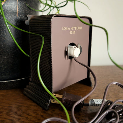 The back of a clock frame with a light and cord attached to the back acrylic piece. Engraved at the bottom 'ASHLEY HAY DESIGN 2026'. It is sitting on a table against a white and plant filled background. 