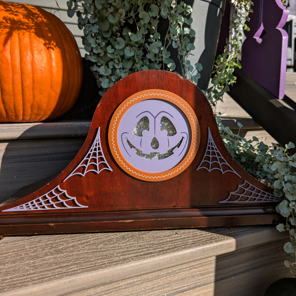 A vintage wood clock frame that has a smiling jack-o-lantern for the face and is sitting on a step against a plants and pumpkins. Matching Spider webs have been added to the front of the frame as well.