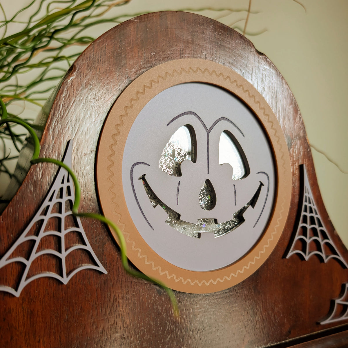 The closeup view of an acrylic grinning jack-o-lantern face inside a vintage wooden clock frame sitting on a table against a white and plant filled background.