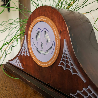 A Halloween-themed wooden vintage mantel clock frame with an acrylic jack-o'-lantern face and spiderweb motifs and is sitting on a table against a white and plant filled background.