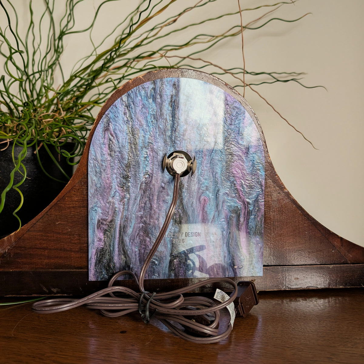 The back of a wooden clock frame with a light and cord attached to the back acrylic piece. Engraved at the bottom 'ASHLEY HAY DESIGN 2026'.  It is sitting on a table against a white and plant filled background.