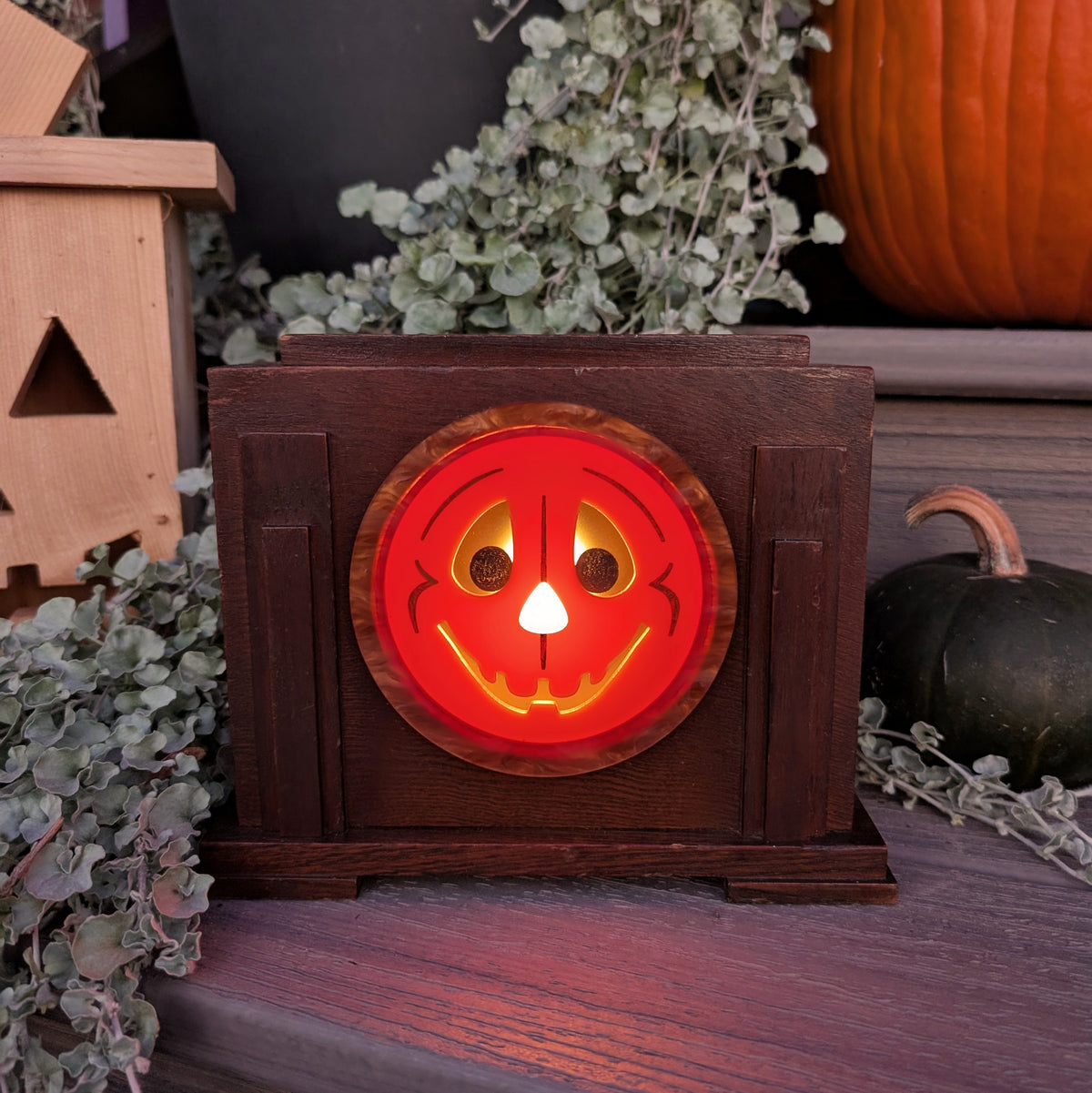 A vintage art deco wood clock frame that has a smiling jack-o-lantern for the face and is sitting on a step against a plants and pumpkins  The scene is dark with the face lit up from inside.