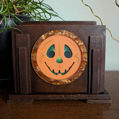 An acrylic smiling jack-o-lantern face inside a vintage wooden clock frame sitting on a table against a white and plant filled background.