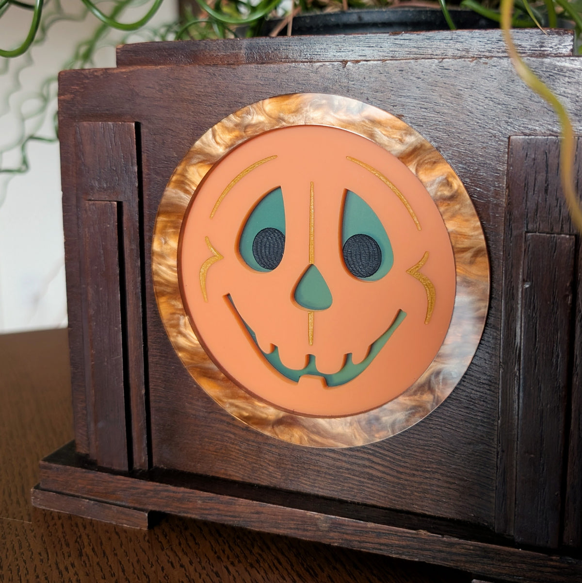 The closeup view of an acrylic smiling jack-o-lantern face inside a vintage wooden clock frame sitting on a table against a white and plant filled background.