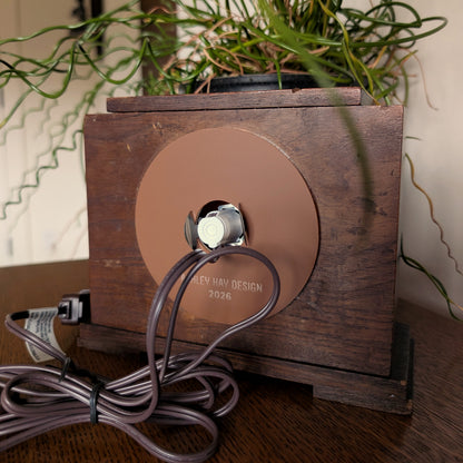 The back of a wooden clock frame with a light and cord attached to the back acrylic piece. Engraved at the bottom 'ASHLEY HAY DESIGN 2026'. It is sitting on a table against a white and plant filled background. 