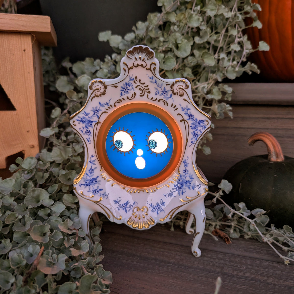 A vintage Victorian-styled ceramic clock frame that has a surprised jack-o-lantern for the face and is sitting on a step against a plants and pumpkins  The scene is dark with the face lit up from inside.