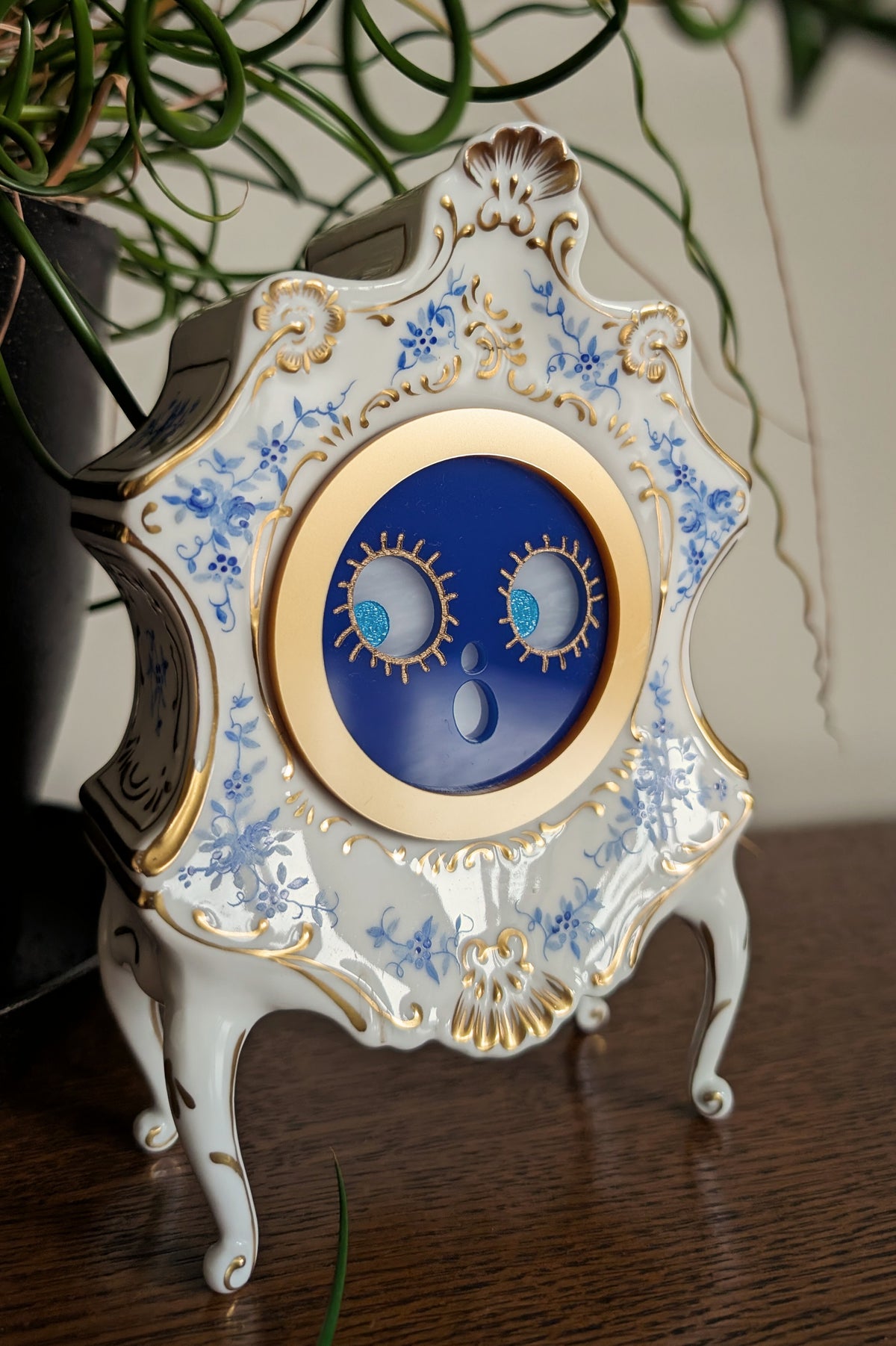 A Victorian-style ceramic clock frame that has acrylic surprised jack-o-lantern face and legs that stand on a table against a white and plant filled background.