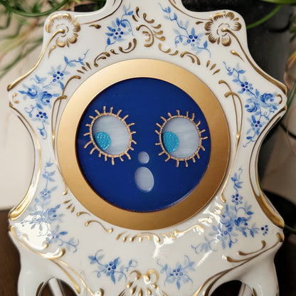 The closeup view of an acrylic smiling jack-o-lantern face inside a vintage ceramic clock frame sitting on a table against a white and plant filled background.
