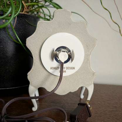The back of a ceramic clock frame with a light and cord attached to the back acrylic piece. Engraved at the bottom 'ASHLEY HAY DESIGN 2026'. It is sitting on a table against a white and plant filled background.