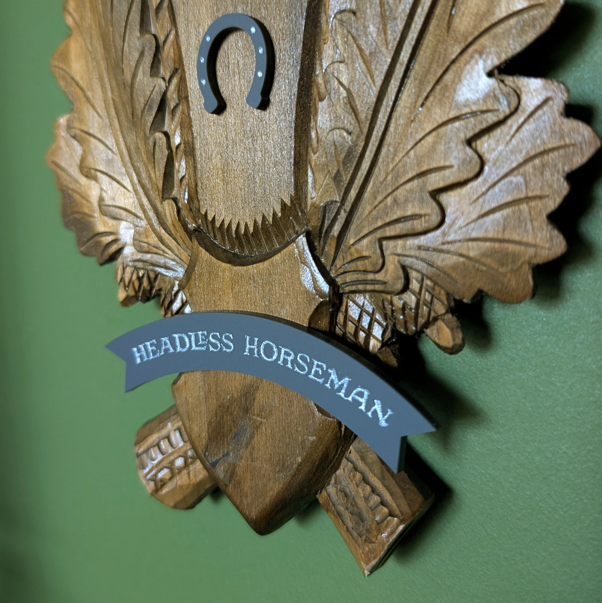 A closeup of the gray banner at the bottom of the carved wood plaque reads 'HEADLESS HORSEMAN' and a small horseshoe sits above it.
