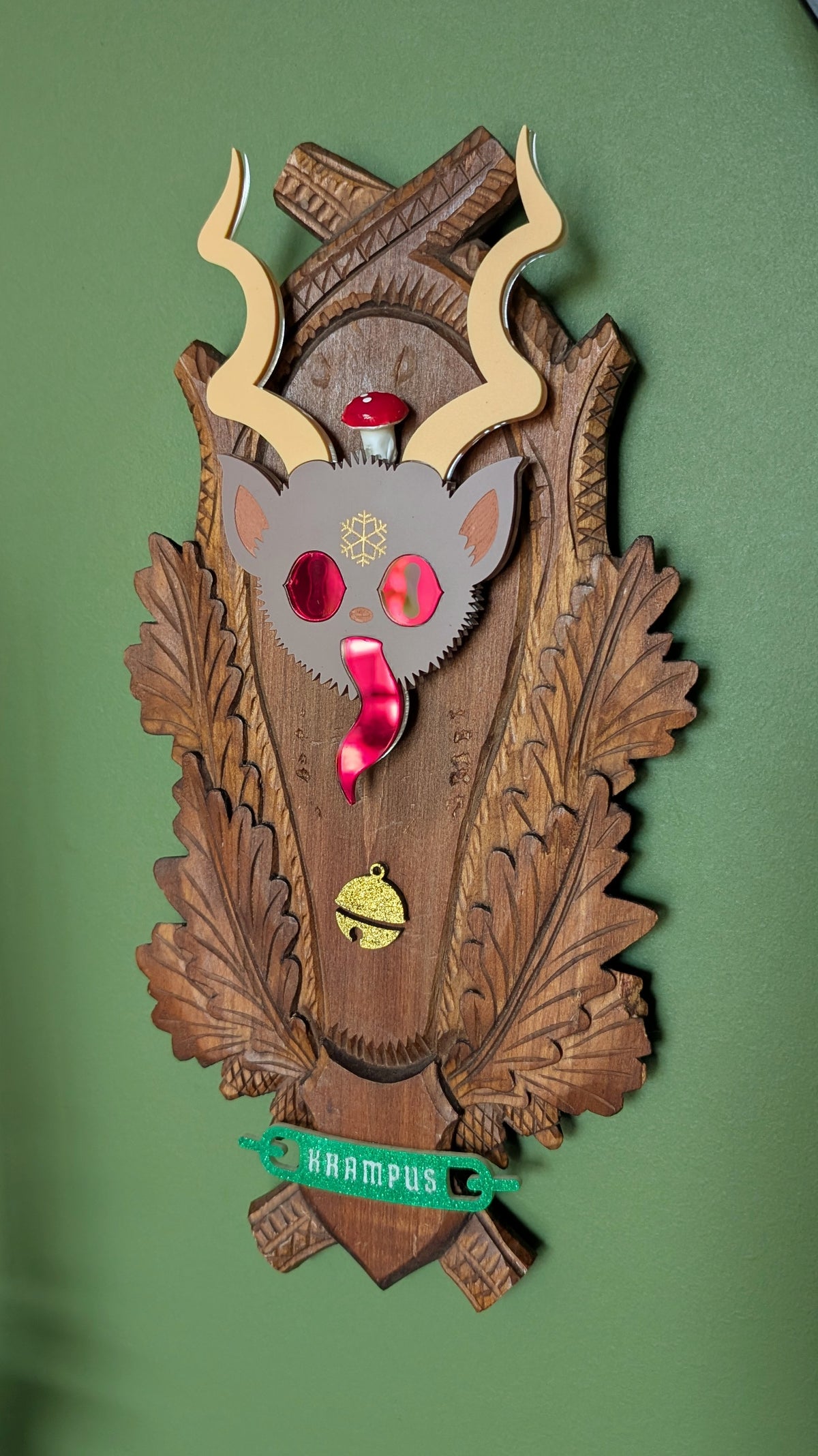 A vintage wooden carved wall plaque featuring an acrylic Krampus head with red mirror eyes and tongue, a vintage mushroom, and a gold bell accent. A green glitter banner at the bottom reads 'KRAMPUS.