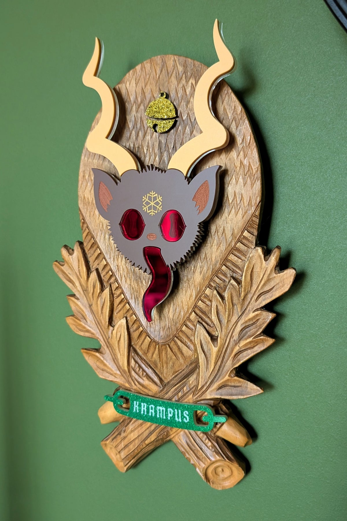 A vintage wooden carved wall plaque featuring an acrylic Krampus head with red mirror eyes and tongue and a gold bell accent. A green glitter banner at the bottom reads 'KRAMPUS'
