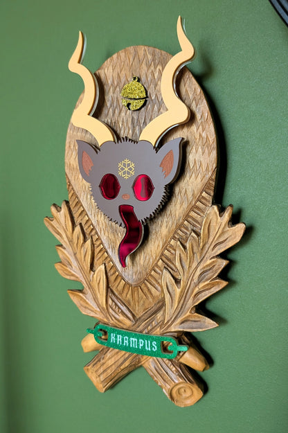 A vintage wooden carved wall plaque featuring an acrylic Krampus head with red mirror eyes and tongue and a gold bell accent. A green glitter banner at the bottom reads 'KRAMPUS'