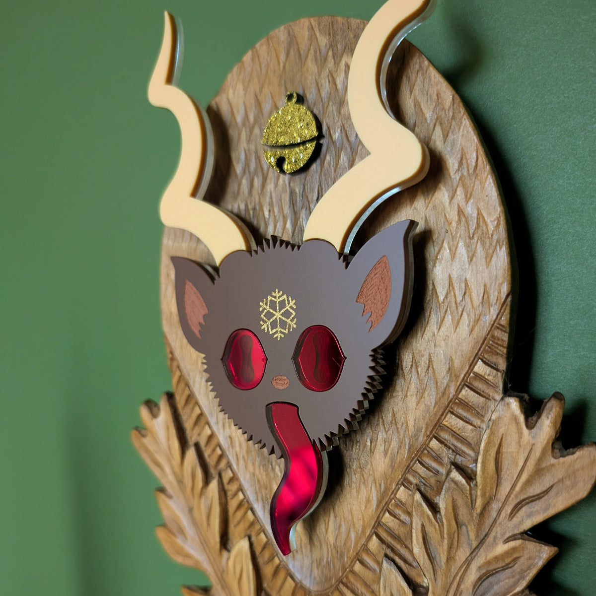 The closeup of a vintage wooden carved wall plaque featuring an acrylic Krampus head with red mirror eyes and tongue and a gold bell accent. 