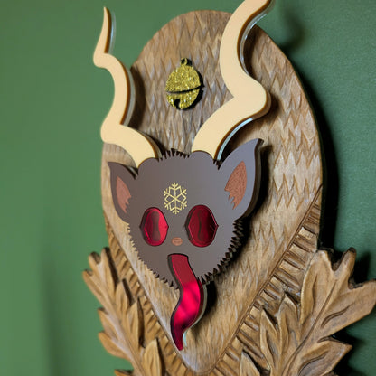 The closeup of a vintage wooden carved wall plaque featuring an acrylic Krampus head with red mirror eyes and tongue and a gold bell accent. 