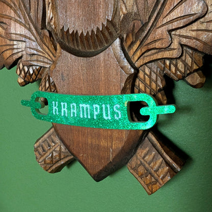 A closeup of the green glitter banner at the bottom of the carved wood plaque reads 'KRAMPUS.