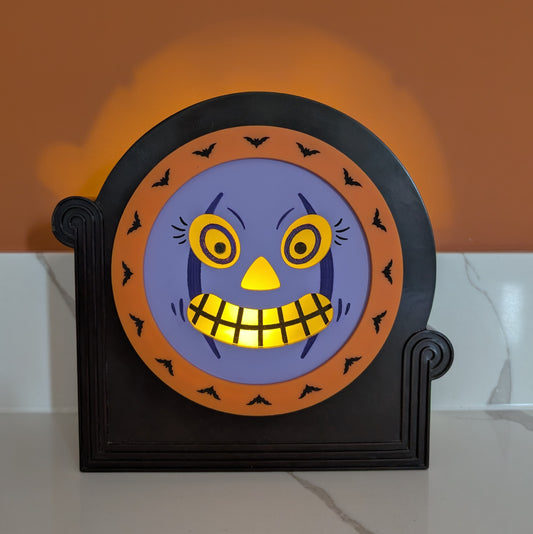 A vintage plastic clock frame repurposed with a purple jack-o-lantern face. The face is lit up.