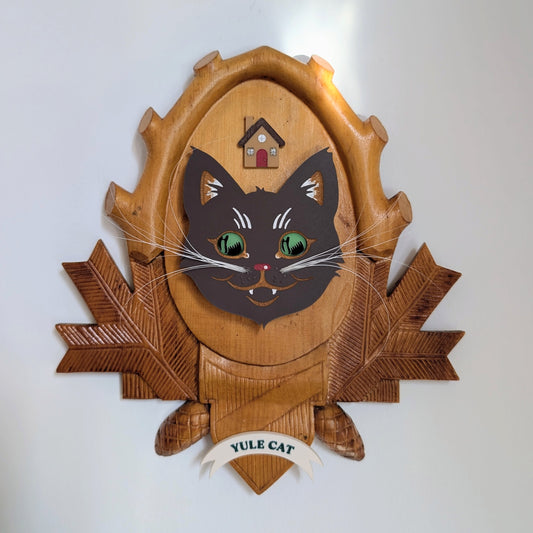 An acrylic Yule Cat head mounted on a wooden plaque hangs on a white wall.
